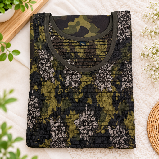 A folded long-sleeve top with a smocked texture, featuring a green and black camouflage pattern with white floral motifs. The garment has a solid olive green crew neck and is displayed on a soft white textured blanket with decorative plants and a woven mat.
