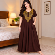 A woman stands barefoot in a bedroom wearing a long brown maxi dress with gold embroidered detailing around the neckline and short sleeves. The dress has side pockets and is shown in a warm, well-lit bedroom setting.